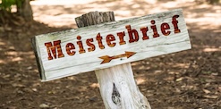 Auf einem Holzpflock ist eine Holztafel befestigt, auf der "Meisterbrief" steht, darunter zeigt ein Pfeil nach rechts
